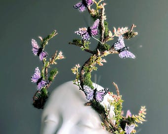 May include: A white mannequin head adorned with a whimsical headpiece. The headpiece features a branch-like structure with moss, small flowers, and several purple butterflies. The Masquerade Store logo is visible at the bottom.