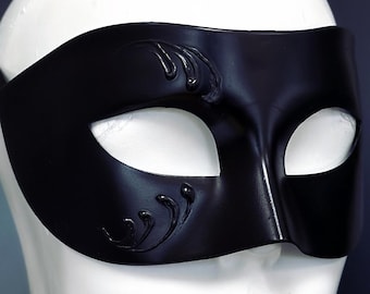 May include: A black masquerade mask with decorative flourishes on the upper left side. The mask has eye openings and a strap to secure it. The mask is displayed on a white mannequin head against a dark background.