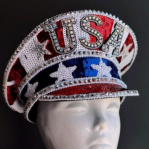 May include: A patriotic, sequined hat with the word "USA" spelled out in sparkling rhinestones. The hat features red, white, and blue colors, with stars and stripes design elements. The hat is displayed on a white mannequin head.