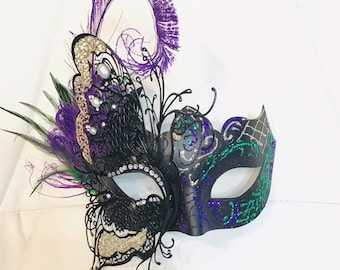 Può includere: Maschera da ballo nera con un design a farfalla. La maschera ha accenti di glitter verde e viola e una piuma.