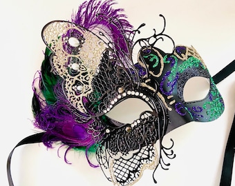 Può includere: Una maschera da carnevale nera con un design a farfalla. La maschera ha dettagli di glitter verde e oro e piume viola.