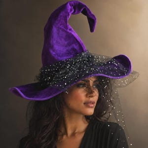May include: A pointed purple velvet hat featuring a wide brim and a black tulle veil overlay. The sheer black tulle is adorned with small, reflective silver sequins that catch the light, creating a shimmering effect across the hat's base.