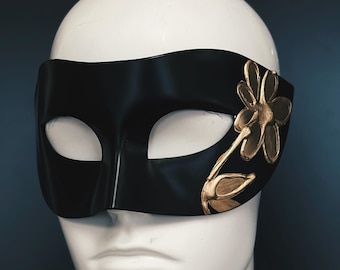 Könnte beinhalten: Eine schwarze Augenmaske mit einem goldenen Blumendesign auf der rechten Seite. Die Maske wird auf einem weißen Schaufensterpuppenkopf vor dunklem Hintergrund präsentiert. Die Maske besteht aus einem glatten, glänzenden Material.