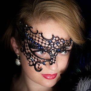 May include: A woman wearing a black lace masquerade mask with blue rhinestones. The mask covers her right eye and part of her forehead. She is wearing a black top and has blonde hair.