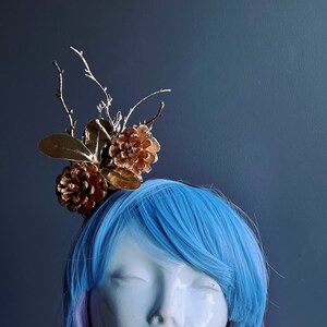 May include: A gold and brown pine cone hairpiece with twigs and leaves. The hairpiece is worn on a mannequin with blue and pink hair.