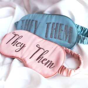 May include: Two satin sleep masks, one pink and one blue, with the words "They Them" embroidered in glitter on each.