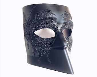 Könnte beinhalten: Eine schwarze Maskenballmaske mit glitzernder Oberfläche und einem einzigartigen, eckigen Design. Die Maske hat ein einzelnes Augenloch und ein spitzes Kinn.