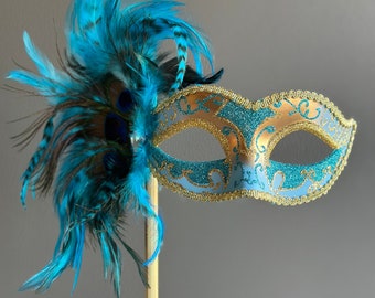 Può includere: Una maschera da carnevale blu e oro con un boa di piume blu attaccato a un bastone dorato.