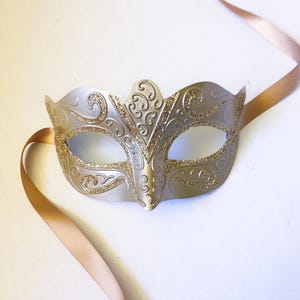 May include: A silver masquerade mask with gold glitter accents and a delicate, ornate design. The mask has a satin ribbon attached for tying.