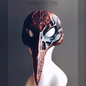Pu&ograve; includere: Una maschera per il ballo in maschera personalizzata, di colore marrone scuro e nero, con un lungo becco appuntito. La maschera presenta un design dettagliato con un motivo circolare sulla fronte e un design a forma di teschio sul becco. La maschera &egrave; esposta su una testa di manichino bianca.