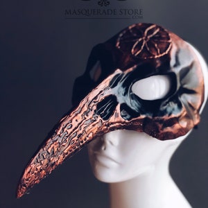 Pu&ograve; includere: Una maschera per il carnevale realizzata su misura con un lungo becco ricurvo color rame e un design a forma di teschio. La maschera ha un'orbita oculare nera e un design floreale color rame nella parte superiore. La maschera &egrave; su una testa di manichino bianca. Il testo "Custom Witche's Mark!" &egrave; nella parte inferiore dell'immagine.