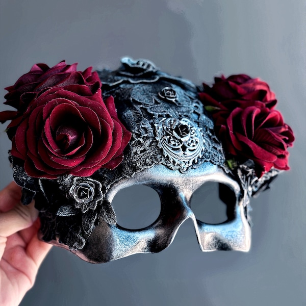 May include: A silver skull masquerade mask with black lace detailing and three burgundy roses on the top.