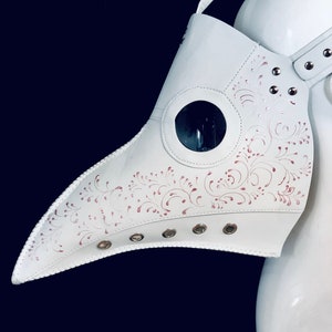 Puede incluir: M&aacute;scara de m&eacute;dico de la peste de cuero blanco con un pico largo y un dise&ntilde;o floral rosa. La m&aacute;scara tiene una pieza ocular transparente y est&aacute; decorada con remaches met&aacute;licos.