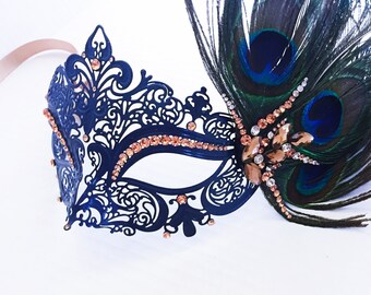May include: Black masquerade mask with intricate cutouts and rhinestones, adorned with a peacock feather plume. The mask has a delicate, ornate design and is accented with gold-toned rhinestones.