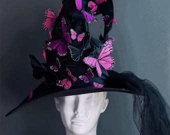 Puede incluir: Un sombrero de bruja negro adornado con mariposas rosas y moradas. El sombrero tiene un ala ancha y una corona alta y puntiaguda. Un velo de tul negro cae por la espalda. El sombrero se exhibe en una cabeza de maniquí blanca.