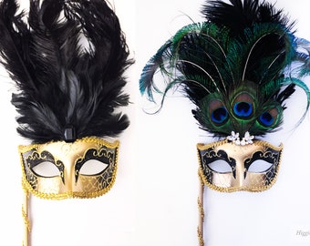 May include: Two gold masquerade masks with black and gold detailing. One mask has black feathers and the other has green and blue peacock feathers.