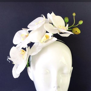 May include: White orchid headband with a black elastic band. The headband is decorated with white orchids and green leaves.