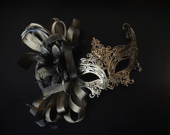 May include: A gold masquerade mask with intricate details and rhinestones. The mask is paired with a black and silver feather and ribbon embellishment.