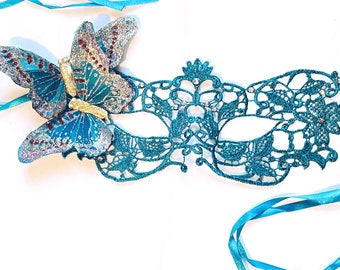 May include: A teal blue masquerade mask with a delicate lace design and a glittery butterfly embellishment. The mask has two teal blue satin ribbons attached.
