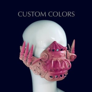 May include: A pink and tan steampunk mask with detailed gear designs and a textured surface. The mask has tubes, spikes, and a central dome. The text "CUSTOM COLORS" is displayed above the mask.