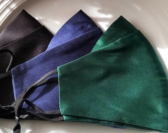 Op de afbeelding: Vier gezichtsmaskers van satijn in zwart, donkerblauw en groen. De maskers zijn gevouwen en op een wit bord gelegd.