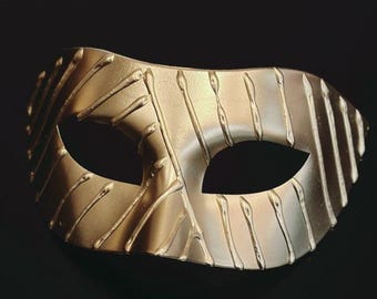May include: A gold-colored masquerade mask with a textured surface and eye openings. The mask has vertical and curved lines, creating a unique design. The mask is set against a black background.