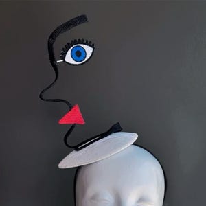 May include: A whimsical headpiece featuring a stylized face design. The black outline of a face is adorned with a blue eye, a red triangle for a mouth, and a small white hat. The headpiece is attached to a black headband.