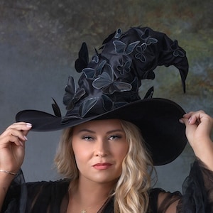 May include: A woman wearing a black dress and a large black witch hat with black butterfly embellishments. The hat is tilted on her head and she is holding the brim with both hands. The background is a dark green and grey textured backdrop.