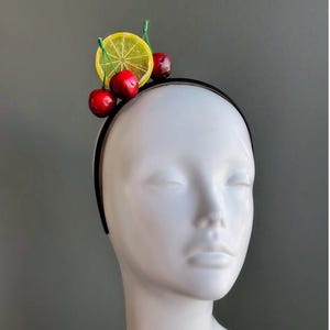 May include: A black headband adorned with a lemon slice and three red cherries. The fruit-themed accessory is displayed on a white mannequin head against a gray background. This playful headband is perfect for themed parties or everyday wear.