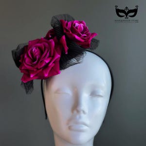 May include: A black headband adorned with three vibrant fuchsia roses and black netting. The roses are the focal point, adding a dramatic flair to the headpiece. The headpiece is displayed on a white mannequin head.