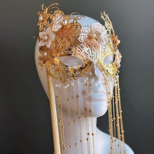 May include: Gold-colored masquerade mask with intricate floral and filigree designs. The mask features dangling chains with small beads and is adorned with white flowers and pearls. The mask is displayed on a white mannequin head.