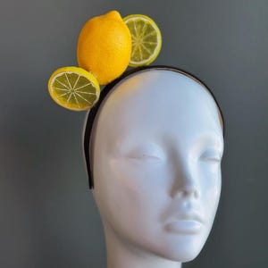 May include: A black headband adorned with a whole yellow lemon and two lemon slices. The fruit-themed headpiece is set against a neutral background, creating a striking contrast. The lemon slices have a detailed, realistic appearance.
