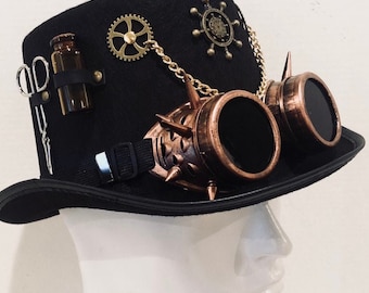 Op de afbeelding: Een zwarte hoge hoed versierd met koperkleurige goggles met stekels, een klein bruin glazen flesje, zilverkleurige scharen, een goudkleurig tandwiel en een scheepsroer-embleem verbonden door een gouden ketting. De hoed staat op een wit etalagehoofd.