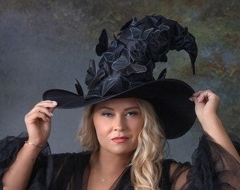 Könnte beinhalten: Eine Frau in einem schwarzen Kleid und einem großen schwarzen Hexenhut mit Schmetterlingsverzierungen. Der Hut ist hoch und spitz, und die Schmetterlinge sind in einem wirbelnden Muster angeordnet. Die Frau schaut mit einem ernsten Ausdruck in die Kamera.