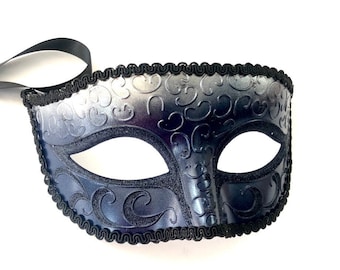 Può includere: Una maschera da ballo blu scuro con un nastro nero. La maschera presenta un motivo decorativo a spirale ed è rifinita con finiture nere. Le aperture per gli occhi sono di forma ovale. La maschera è posta su uno sfondo bianco.