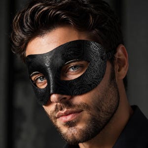 May include: A black masquerade mask featuring an intricate embossed paisley pattern across the surface. The mask covers the eye area and is secured with a thin black strap on the side, showcasing a detailed texture and a matte finish throughout.