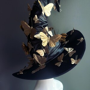 May include: A black witch hat adorned with numerous gold-colored butterfly decorations. The hat has a wide brim and a tall, pointed crown. The butterflies are of varying sizes and are attached to the hat with thin wires. The hat is displayed on a white mannequin head.