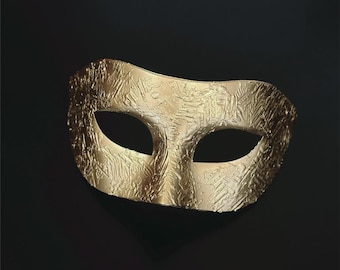 May include: A gold-colored masquerade mask with a textured surface. The mask has two eye openings and is set against a black background. The mask is likely used for costume parties or theatrical performances.