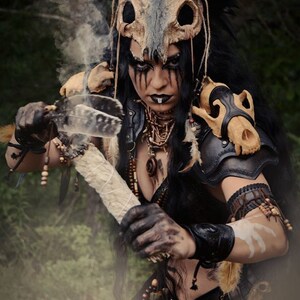 May include: A woman wearing a costume with a skull headdress and leather armor holds a smoldering stick with feathers. She has dark hair, dark eyes, and dark makeup.