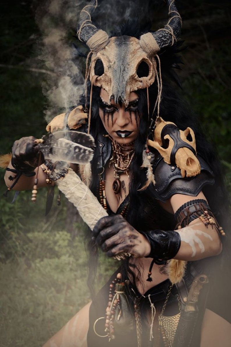 May include: A woman wearing a costume with a skull headdress and leather armor holds a smoldering stick with feathers. She has dark hair, dark eyes, and dark makeup.