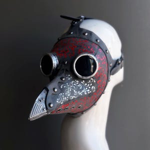 May include: A black and red plague doctor mask with silver rivets and a long, beaked nose. The mask features round eye lenses and is decorated with a red floral pattern. The mask is displayed on a white mannequin head.