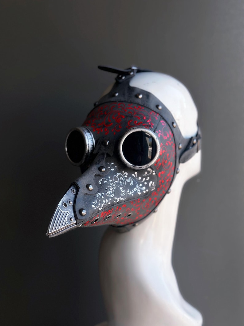 May include: A black and red plague doctor mask with silver rivets and a long, beaked nose. The mask features round eye lenses and is decorated with a red floral pattern. The mask is displayed on a white mannequin head.