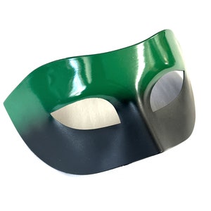 Puede incluir: Una máscara de carnaval verde y negra con un solo agujero para el ojo. La máscara está hecha de un material liso y brillante.