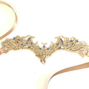 May include: A gold lace headpiece with blue gemstones. The headpiece has a delicate, openwork design with a symmetrical shape. A beige ribbon is attached to the headpiece. The headpiece is on a white background.