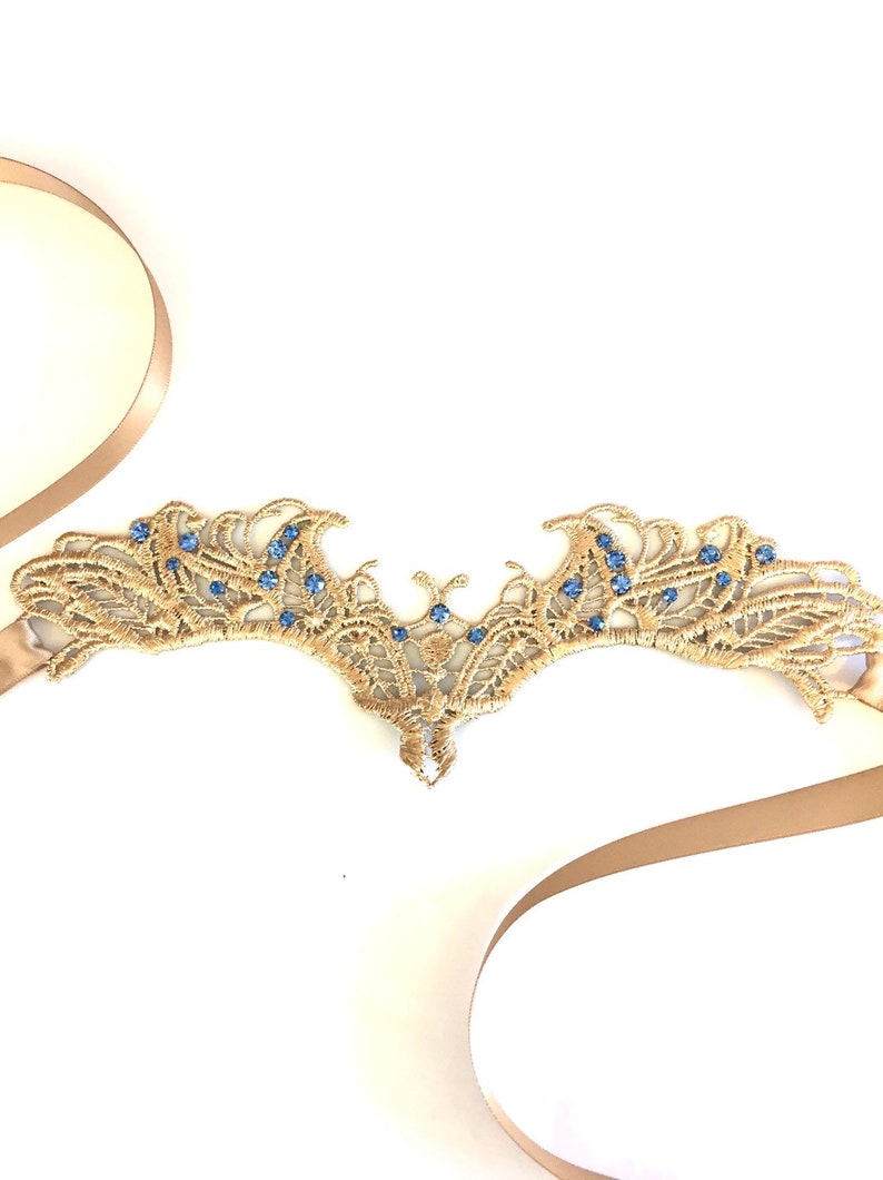 May include: A gold lace headpiece with blue gemstones. The headpiece has a delicate, openwork design with a symmetrical shape. A beige ribbon is attached to the headpiece. The headpiece is on a white background.