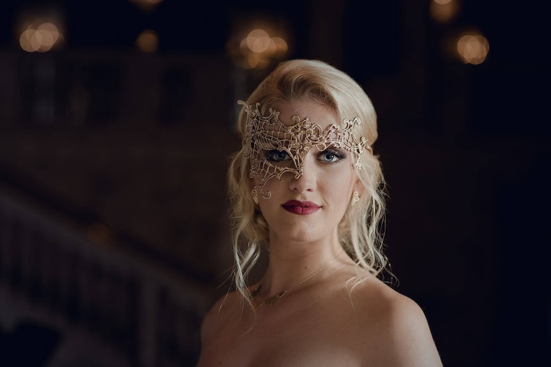 May include: A woman with blonde hair wears a gold lace masquerade mask. She is looking at the camera with a soft smile.