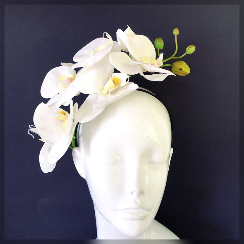 May include: White orchid headband with a black elastic band. The headband is decorated with white orchids and green leaves.