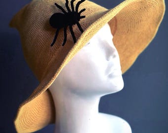 May include: A tan witch hat with a black spider embellishment. The hat is made of a woven material and has a wide brim.