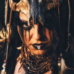 May include: A woman with dark hair and dark makeup wears a skull headdress and a beaded necklace with a pendant.