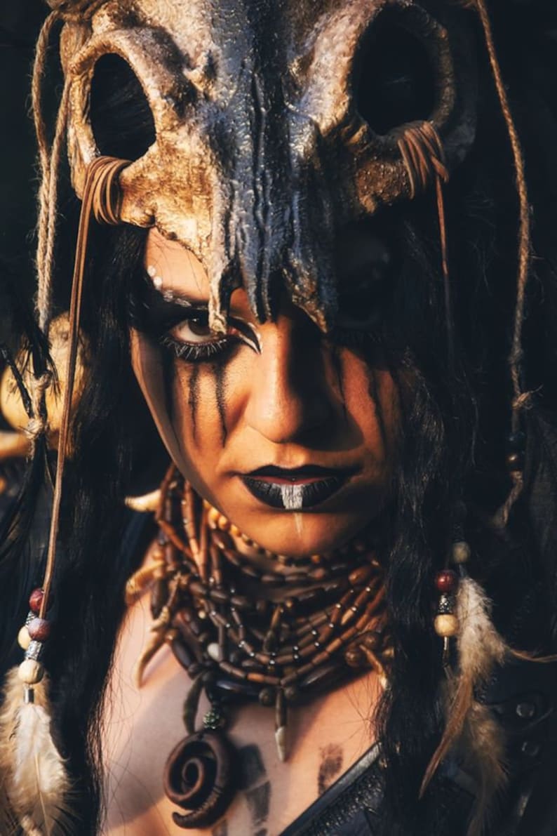 May include: A woman with dark hair and dark makeup wears a skull headdress and a beaded necklace with a pendant.